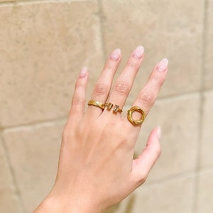 May include: A hand wearing three gold rings. The rings are a thin band, a small ring with two clear stones, and a larger ring with a circular design.
