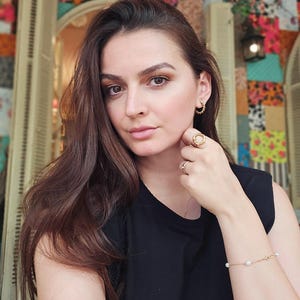 May include: A woman with long brown hair wearing a black sleeveless shirt and gold jewelry. She is wearing a gold bracelet with small white pearls and a gold ring on her finger.