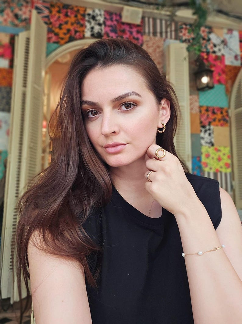 May include: A woman with long brown hair wearing a black sleeveless shirt and gold jewelry. She is wearing a gold bracelet with small white pearls and a gold ring on her finger.