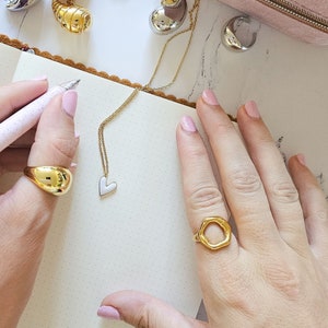 May include: A close-up of two hands wearing gold rings. The ring on the left hand is a solid gold oval shape. The ring on the right hand is a large, open, irregular hexagon shape. The rings are on a white background.