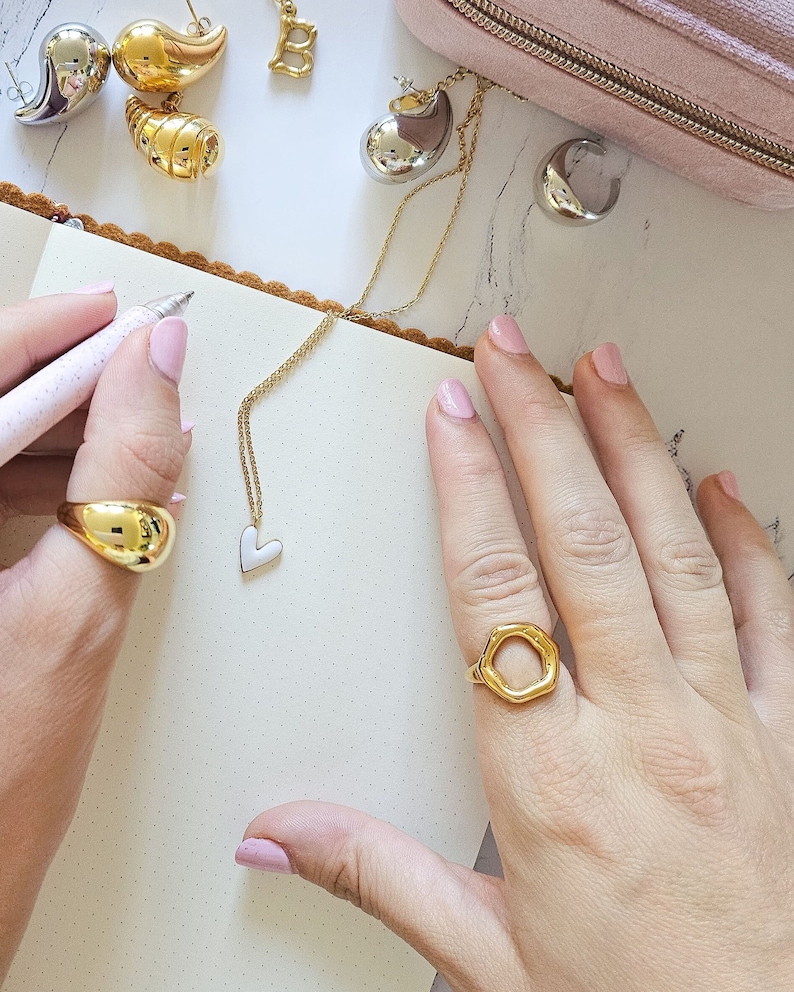 May include: A close-up of two hands wearing gold rings. The ring on the left hand is a solid gold oval shape. The ring on the right hand is a large, open, irregular hexagon shape. The rings are on a white background.