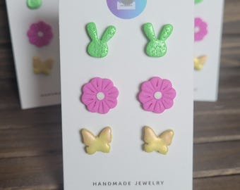 Puede incluir: Un conjunto de seis pendientes hechos a mano con un diseño de conejo verde, flor rosa y mariposa amarilla. Los pendientes se muestran en una tarjeta blanca con el texto "HANDMADE JEWELRY".