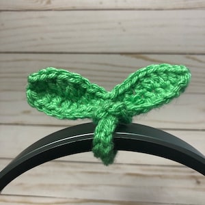 May include: Green crocheted leaf headband for headphones. The headband is made of green yarn and is shaped like a leaf. It is designed to be placed over the headband of headphones.