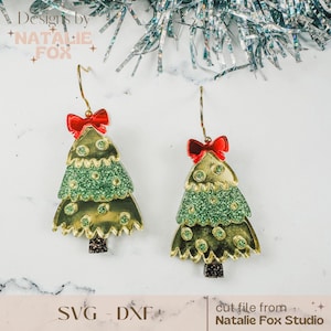 Puede incluir: Un par de pendientes de árbol de Navidad dorados con purpurina verde y lazos rojos. Los pendientes están sobre un fondo blanco con una guirnalda plateada.