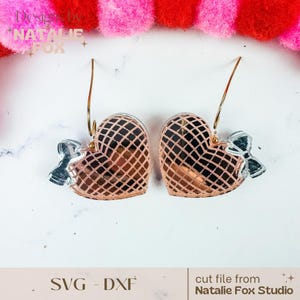 May include: A pair of gold heart-shaped earrings with a delicate mesh pattern and a small silver bow on each earring. The earrings are on a white surface.