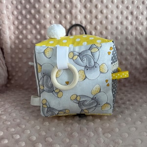 May include: A yellow and gray fabric cube with a white pom-pom on top. The cube has a white ring attached to it and is decorated with gray elephants and yellow polka dots.