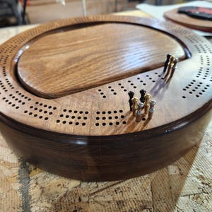 Op de afbeelding: Een houten cribbagebord met een donkerbruine buitenkant en een lichtere bruine binnenkant. Het bord heeft een cirkelvormig ontwerp met een centraal speelveld en talrijke gaten voor pinnen. Verschillende pinnen in zwart, goud en zilver zijn in het bord gestoken.