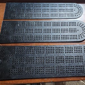 May include: Three black cribbage boards with numerous drilled holes for peg placement. The boards are arranged on a wooden surface, showcasing their distinct shapes and the pattern of the holes. The boards are a dark, matte black.
