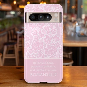 May include: A pink phone case with a white lace pattern. The case has a rectangular design with a section at the bottom that includes the text "Be joyful in hope, patient in affliction, and faithful in prayer. ROMANS 12:12."