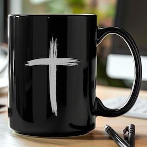 May include: Black ceramic mug with a white cross design. The cross is a simple, brushstroke style, centered on the mug. The mug has a large handle and is sitting on a wooden surface.
