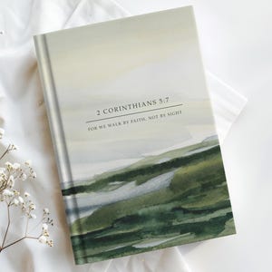 Könnte beinhalten: Ein Hardcover-Journal mit einem Aquarell-Landschaftsdesign in Grün-, Grau- und Gelbtönen. Der Einband trägt den Text "2 Korinther 5,7" und "Denn wir wandeln im Glauben, nicht im Schauen."