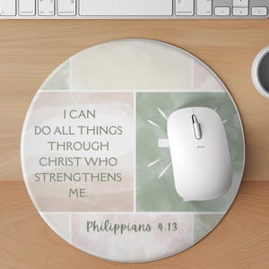 May include: A round mouse pad featuring a watercolor-style background in muted green, beige, and white tones. The text reads, "I CAN DO ALL THINGS THROUGH CHRIST WHO STRENGTHENS ME. Philippians 4:13." A white computer mouse rests on the surface.