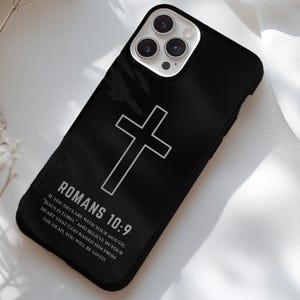 Op de afbeelding: Zwarte telefoonhoes met een wit kruis en de tekst "ROMANS 10:9" met een bijbelvers. De hoes heeft een glanzende afwerking en is ontworpen voor een smartphone.