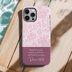 以下が含まれることがあります： ピンクと白のフローラルな携帯電話ケース。ローマ人への手紙12章12節からの引用文「いつも主に喜んでいなさい。繰り返しますが、喜んでいなさい。」が書かれています。引用文は、ワインレッドの背景に白い文字で書かれています。