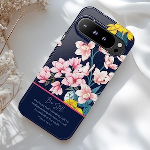 May include: A navy blue phone case featuring a floral design with pink and yellow flowers. The case has the text "Be Still" and a Bible verse printed on it. The case is designed to fit a smartphone.