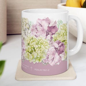 Op de afbeelding: Witte keramische mok met een bloemenontwerp met roze en groene hortensia's. De mok heeft een roze basis met de tekst "PSALM 116:1-2" erop gedrukt.