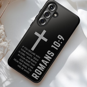 May include: Black phone case with a silver cross and the text "ROMANS 10:9". The case also features a quote from Romans 10:9. The phone case has a triple camera setup.