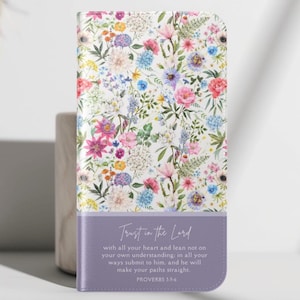Puede incluir: Una funda de teléfono con estampado floral, con una variedad de flores coloridas sobre un fondo blanco. La parte inferior es de color lavanda, con el texto "Trust in the Lord" y un versículo bíblico.