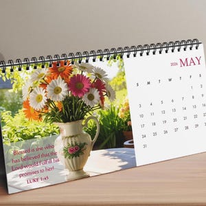 May include: A desk calendar for May 2026, featuring a floral arrangement in a decorative vase. The vase is filled with white, orange, and pink daisies. The calendar includes a Bible verse: Luke 1:45.