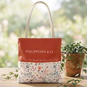 May include: A rectangular tote bag with two off-white shoulder straps. The upper portion is solid burnt orange with the white text PHILIPPIANS 4:13 and the script text I can do all this through him who gives me strength. The lower part features a floral print.