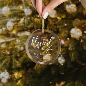 May include: Clear, round Christmas ornament with gold trim and a gold ribbon for hanging. The ornament features the word "blessed" in cursive, along with the phrase "It is more to give than to receive." The text "Jesus of Nazareth, Acts 20:35" is also included.