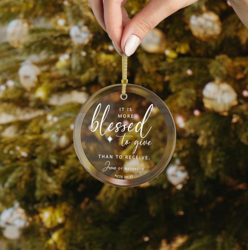 May include: Clear, round Christmas ornament with gold trim and a gold ribbon for hanging. The ornament features the word "blessed" in cursive, along with the phrase "It is more to give than to receive." The text "Jesus of Nazareth, Acts 20:35" is also included.