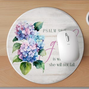 May include: A round mouse mat with a white woodgrain background. A watercolour illustration of purple and blue hydrangeas is on the left side of the mat. The text "PSALM 46:5" and "God is within her, she will not fall." is on the right side of the mat.