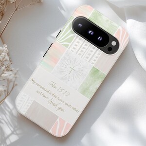 May include: A phone case with a pastel patchwork design in shades of pink, green, and gray. The case features the text "John 15:12" and a quote about love. The phone case has a camera lens and flash.