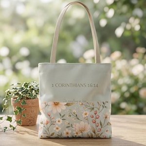 May include: A light sage green tote bag featuring two long shoulder straps. The upper portion displays the text 1 CORINTHIANS 16:14, while the lower section is decorated with a botanical print of various flowers and foliage in soft pastel tones.