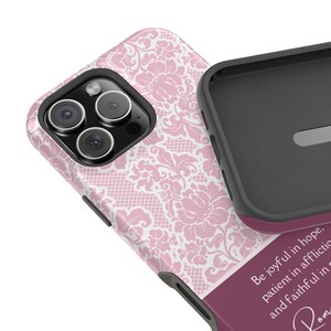 以下が含まれることがあります： ピンクと白のダマスク柄の携帯電話ケース。ケースの下部には、「希望に満ちて喜び、苦難に耐え、祈りに忠実であれ」という引用が書かれています。