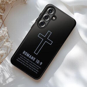 May include: Black phone case featuring a white cross and the text "ROMANS 10:9". The case also includes a quote from Romans 10:9. The phone case has a glossy finish and a camera module.