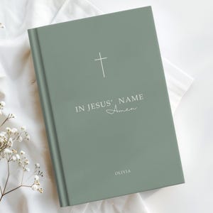 Może przedstawiać: Szałwiowy zielony notatnik z białym krzyżem i napisem "IN JESUS' NAME Amen" oraz imieniem "OLIVIA". Notatnik jest zamknięty i ma minimalistyczny design. Okładka jest w jednolitym kolorze.
