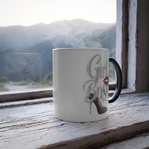 May include: A white ceramic mug with the words "Girl Boss" in a silver, distressed font, a red lipstick kiss, and a black high heel. The mug has a black handle and is steaming.