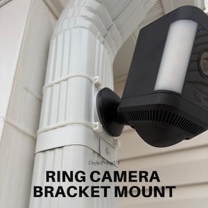 May include: A black Ring camera mounted on a white gutter using a bracket. The camera has a built-in light and is secured with white zip ties. The text "RING CAMERA BRACKET MOUNT" is displayed at the bottom of the image.