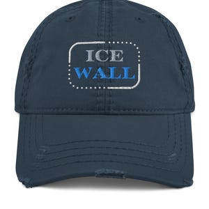 May include: A navy blue baseball cap with a distressed finish. The cap features the embroidered text "ICE WALL" in grey and blue, enclosed within a white dotted rectangle. The cap has a curved peak.