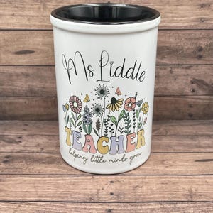 May include: White ceramic utensil holder with a black rim. It has a floral design with the text "Ms. Liddle Teacher helping little minds grow".