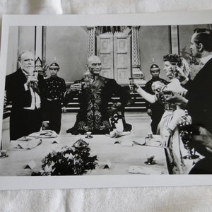 May include: A black and white photo of a group of people seated at a table, raising their glasses in a toast. The people are dressed in formal attire, and the table is set with fine china and silverware.