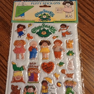 May include: A package of Cabbage Patch Kids puffy stick-ons featuring various illustrated characters, animals, and text including Best Friends, I Love My Cabbage Patch Kid, and Rain Rain Go Away. The stickers are arranged on a white backing card.