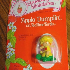 May include: A vintage Strawberryland Miniatures Apple Dumplin' on TeaTime Turtle figure in original packaging. The orange card features green leaves, white flowers, and text including "Scented like apples" and "We're both berry little."