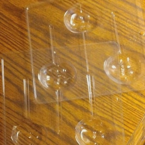 May include: A clear plastic candy mold featuring four circular cavities designed for lollipops. Each circular depression is connected to a narrow, straight channel intended for a stick, arranged in a rectangular tray format with smooth edges.