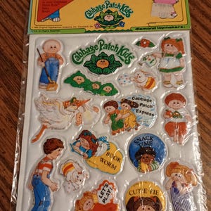 May include: A package of Cabbage Patch Kids Puffy Stick-Ons featuring various stickers of children, animals, and text including Cabbage Patch Express, Book Worm, Snack Time, Let's Play, and Cutie-Pie. The stickers are on a white backing card.