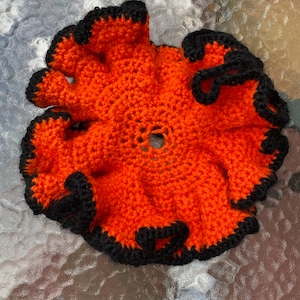May include: Orange crocheted coaster with a black crocheted border. The coaster has a ruffled edge and a hole in the center.