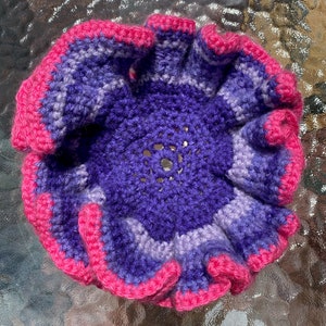 May include: A crocheted flower with a pink and purple ombre design. The flower has a ruffled edge and a center hole.