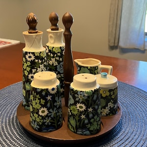 May include: A vintage ceramic spice set with a dark floral pattern, including salt and pepper shakers, a creamer, and a sugar bowl, all on a wooden tray. The set features a dark background with white, blue, and green floral designs.