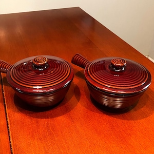 May include: Two brown ceramic bowls with lids and handles. The bowls have a swirl pattern and are on a wooden surface.