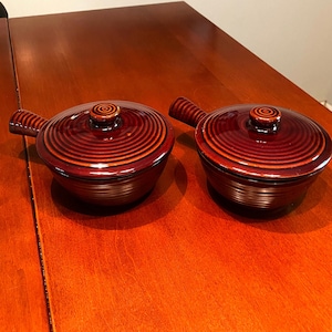 May include: Two brown ceramic bowls with lids and handles. The bowls have a swirl pattern and are on a wooden surface.