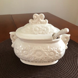 May include: White ceramic soup tureen with a floral design and a lid. The tureen has a matching ladle.