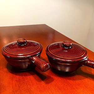 May include: Two brown ceramic ramekins with lids and handles. The ramekins have a grooved design and are sitting on a wooden surface.
