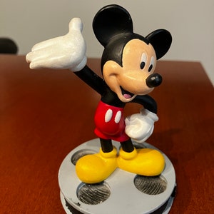 May include: A black and white Mickey Mouse figurine standing on a film reel base. Mickey is wearing red shorts with two black buttons and yellow shoes. He is waving with his right hand.