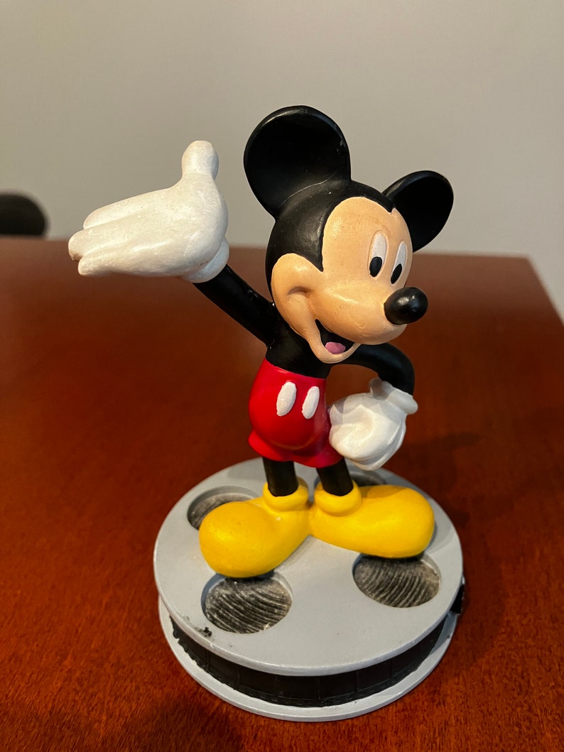 May include: A black and white Mickey Mouse figurine standing on a film reel base. Mickey is wearing red shorts with two black buttons and yellow shoes. He is waving with his right hand.